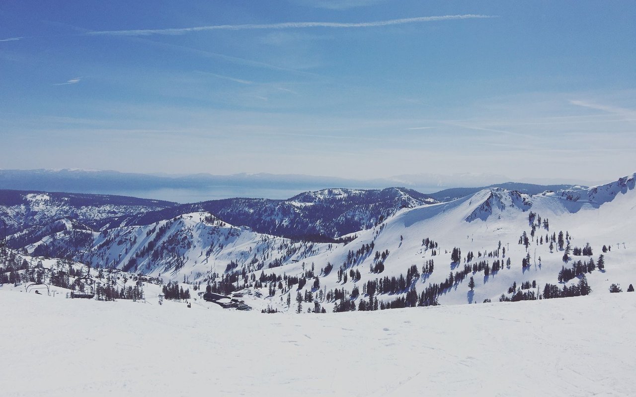 Snow-covered ski slopes and forested ridges overlook Lake Tahoe under a clear blue sky.
