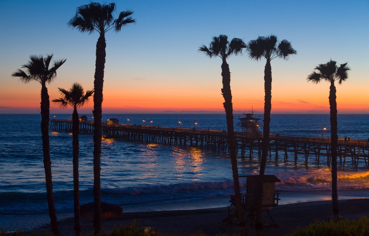 Ocean pier at sunset with silhouetted palm trees and lights along the walkway.