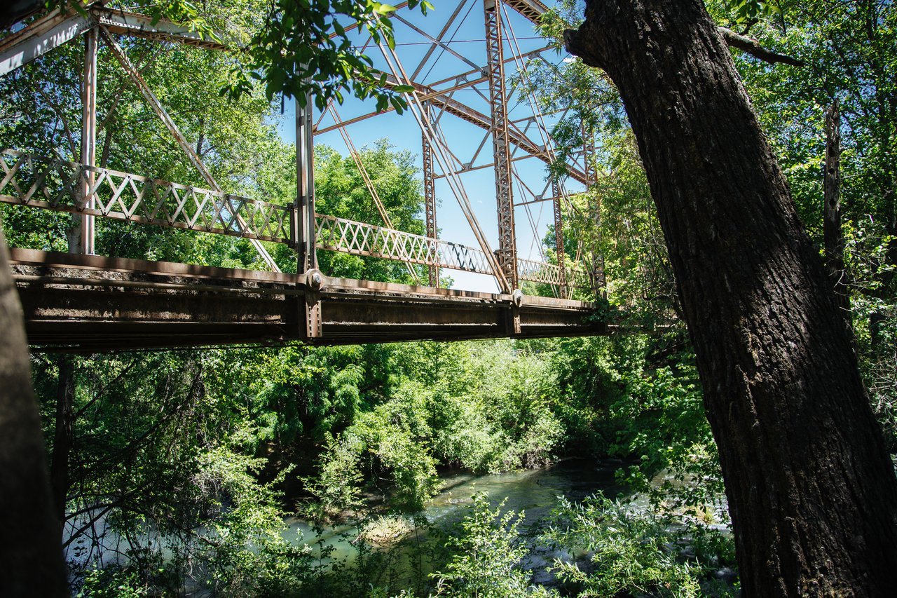 Steel truss bridge crossing a shaded river amid dense green trees.