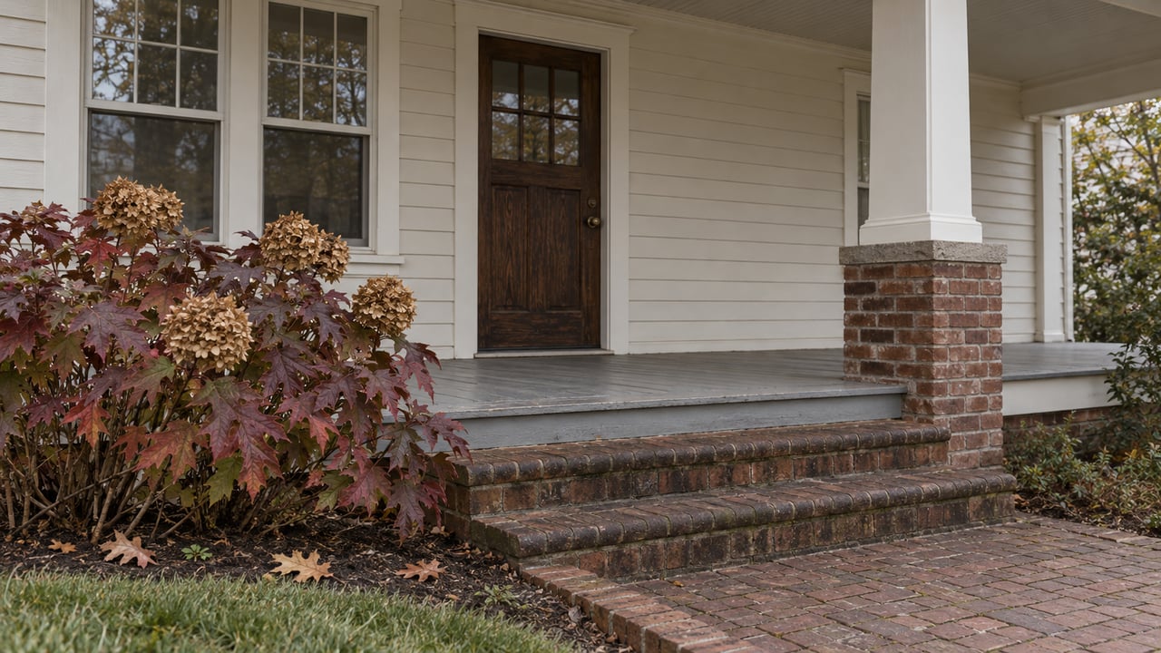 An ivory clapboard home's covered porch, oak front door and weathered brick steps beside burgundy oakleaf hydrangea.