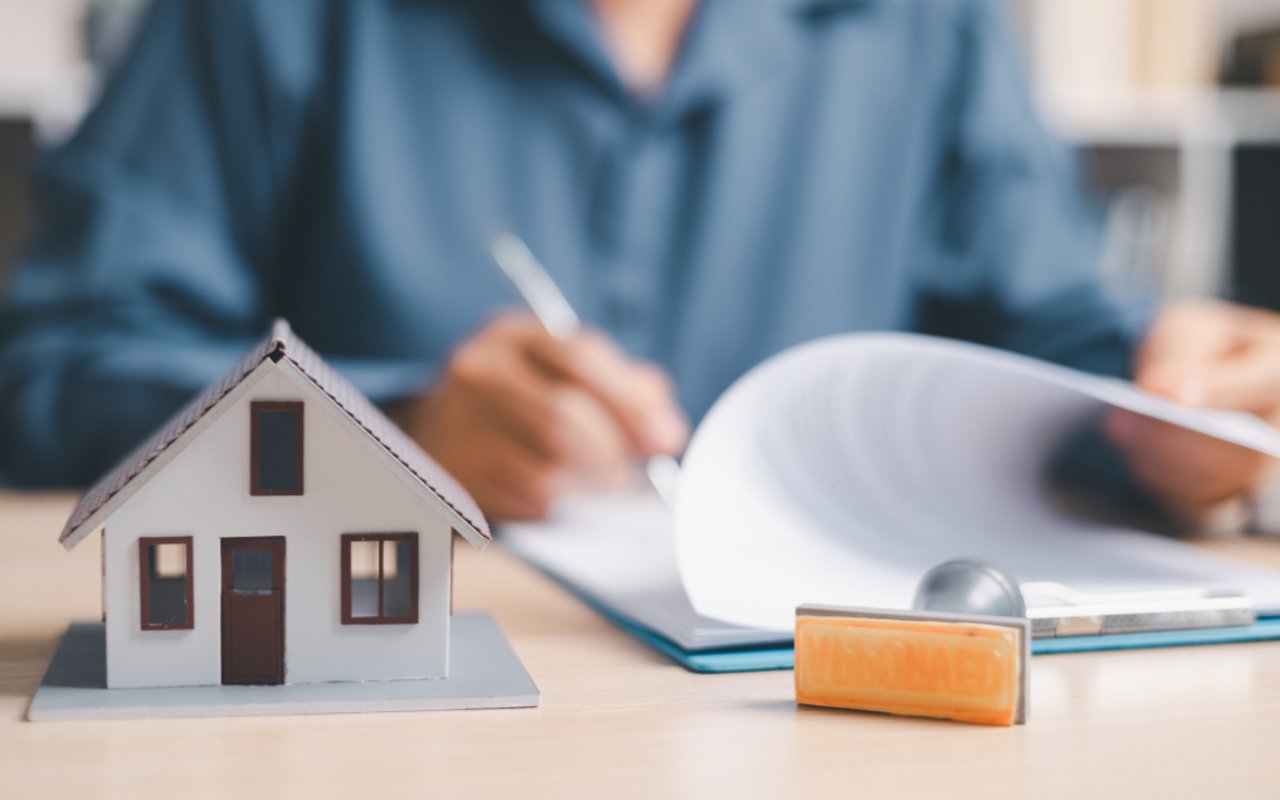 Miniature house beside loan paperwork as someone writes in the background.
