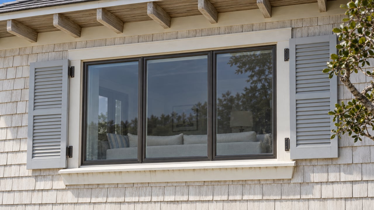 Ivory shingle facade of an elevated home, with a broad bronze-framed window, pale-gray shutters and a weathered cedar eave beside a live oak branch.