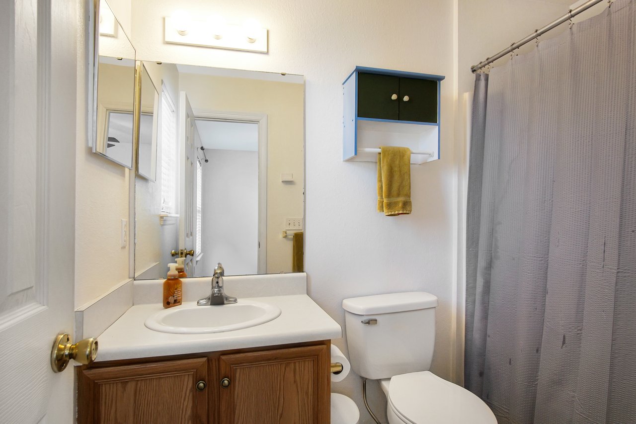 Small bathroom with a wooden cabinet under the sink, large mirror, and light fixture. A towel hangs beneath a wall cabinet next to a gray shower curtain.