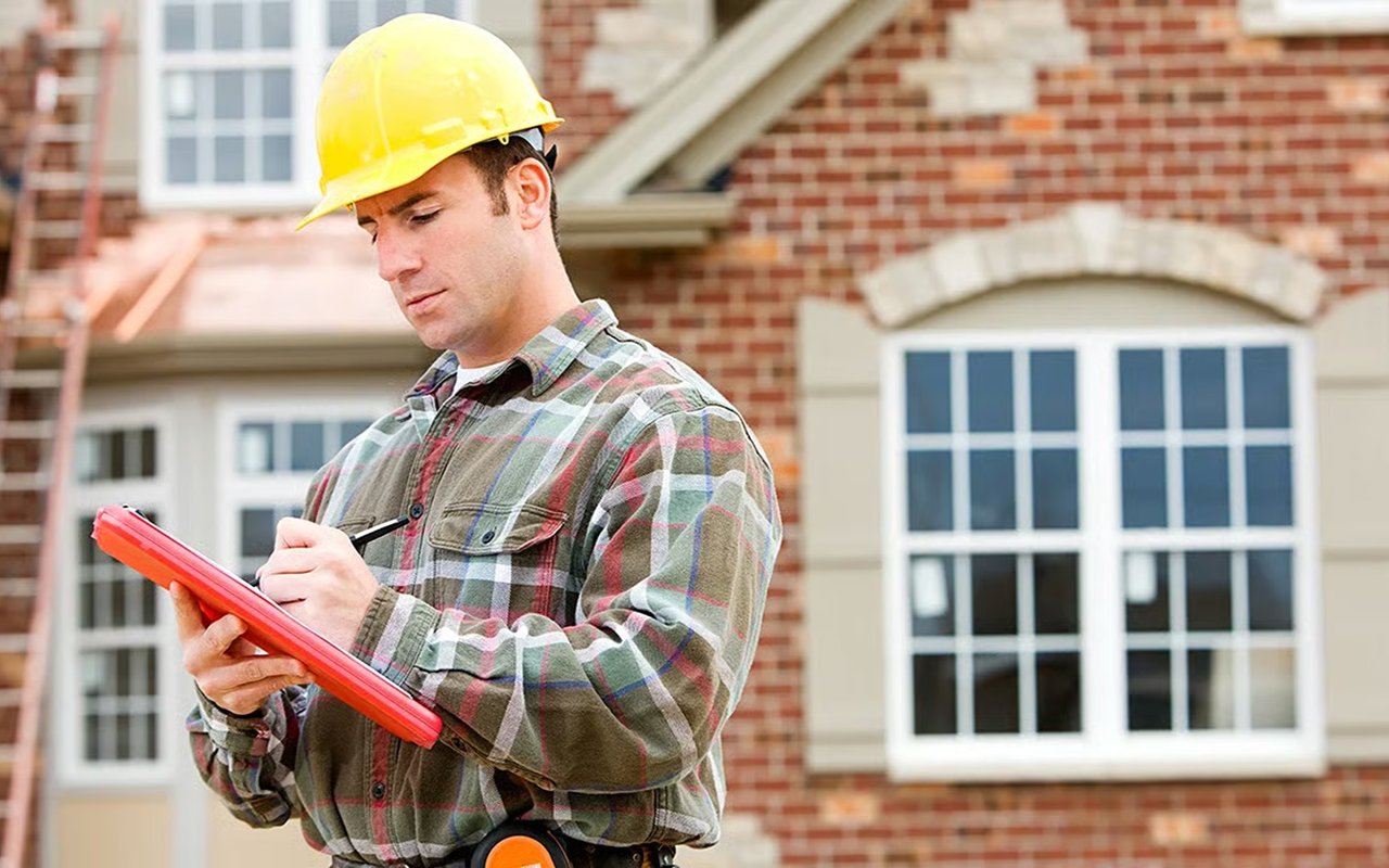 Home inspector in a yellow hard hat records notes on a clipboard outside a brick house.