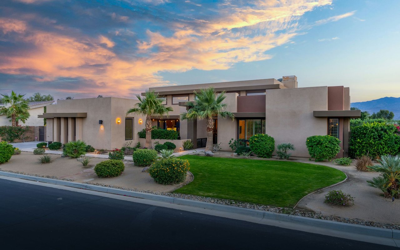 Modern desert home with palm trees, landscaped lawn, and mountains at sunset.