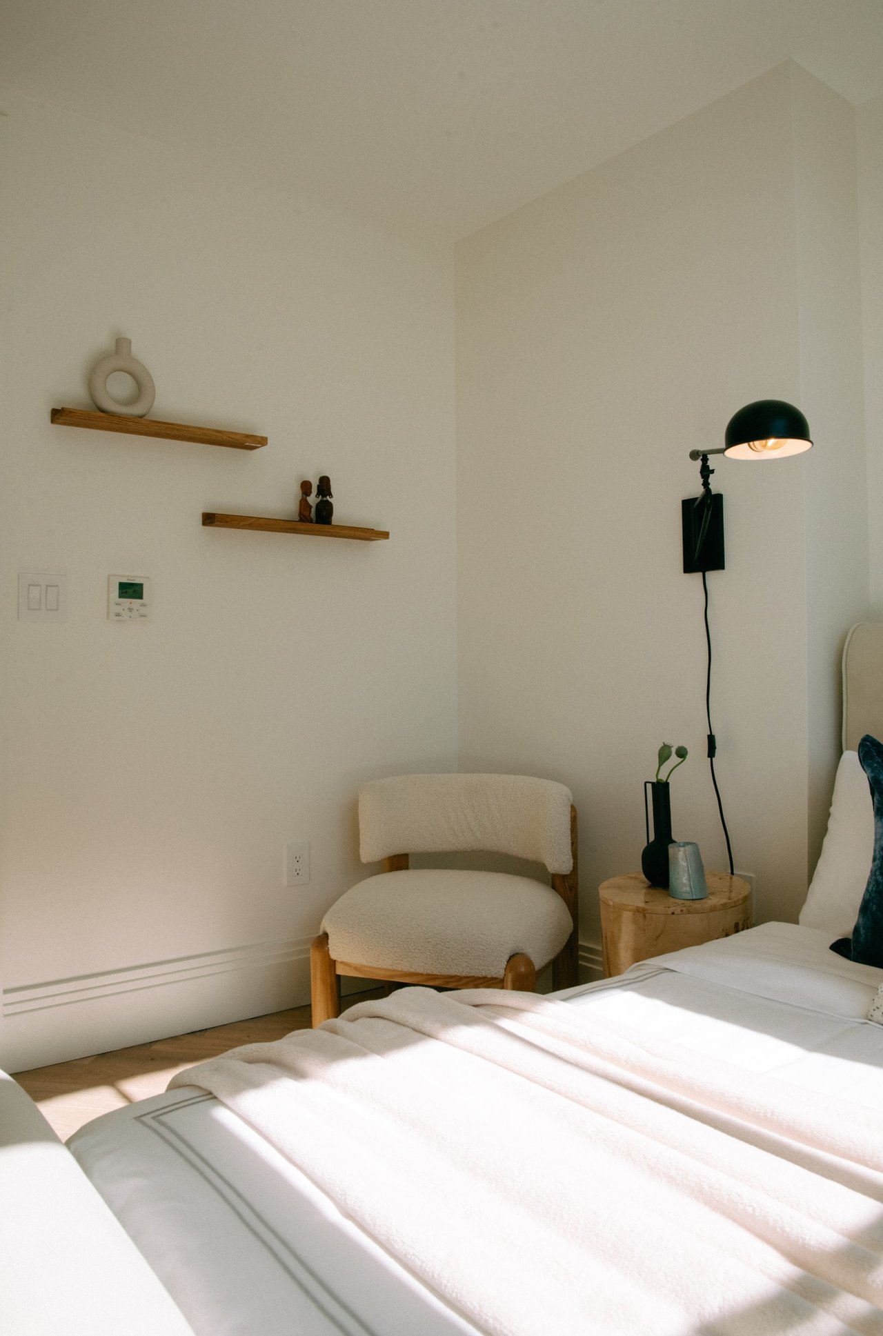 Bedroom with white bedding, a bouclé chair, wooden shelves, and a lit wall lamp.