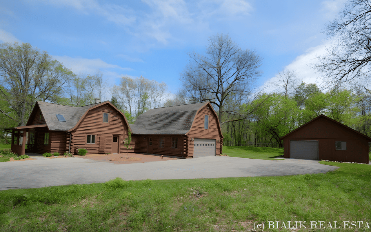 Brown log-style home with an attached garage, separate outbuilding, and wide paved driveway.