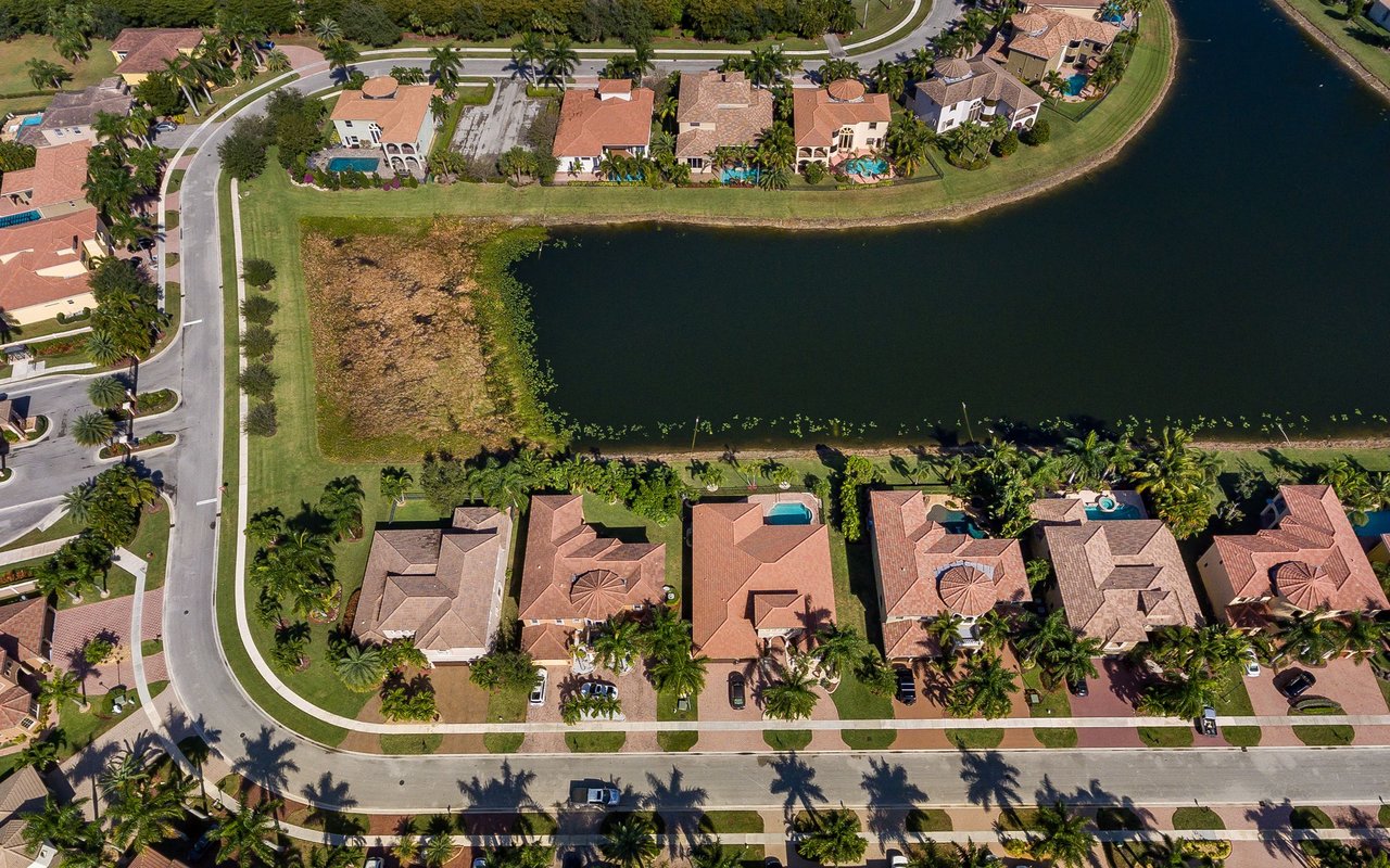 Aerial view of terracotta-roofed homes lining a large lake, with palm trees and swimming pools.