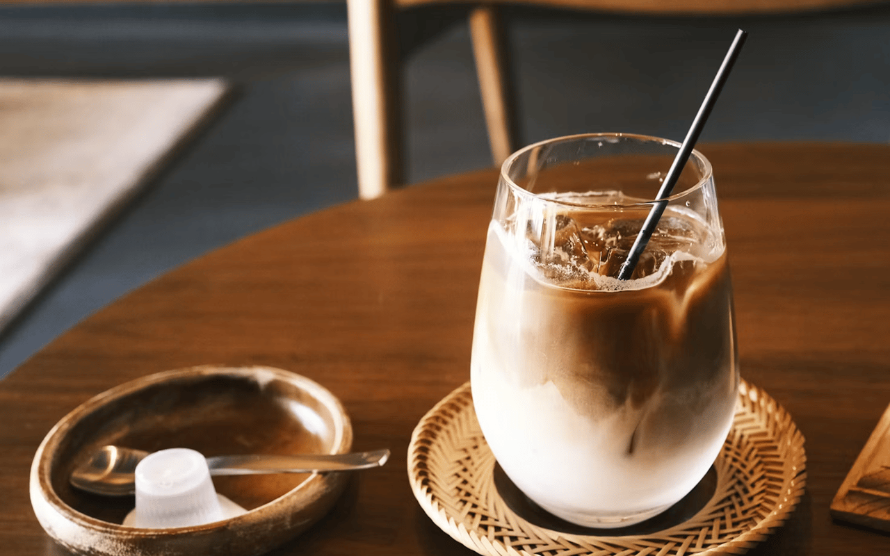 Iced coffee with milk and a black straw on a woven coaster beside a bowl and spoon.