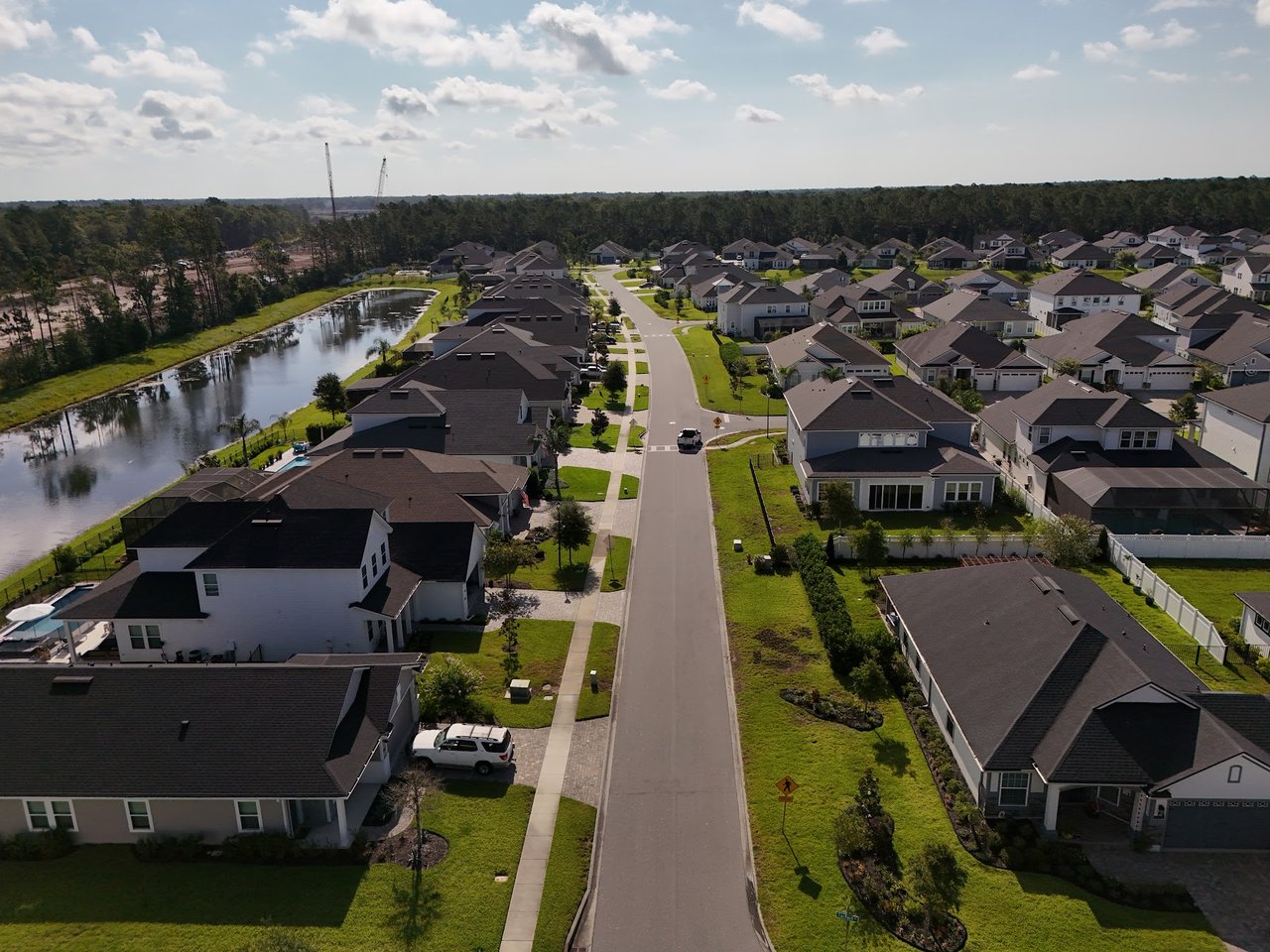 Aerial view of homes lining a neighborhood road beside a pond and wooded area.