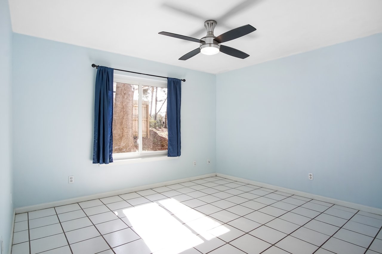 Empty room with light blue walls, white tiled floor, and a ceiling fan. Sunlight streams through a window with blue curtains, creating a calm atmosphere.