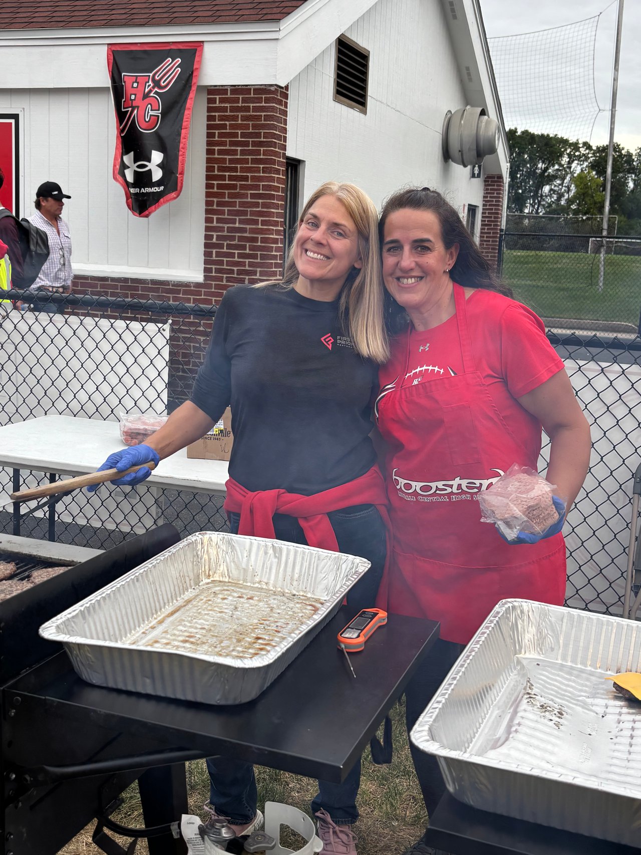 Two women volunteer at a Hinsdale Central football food stand, one wearing a red boosters apron.