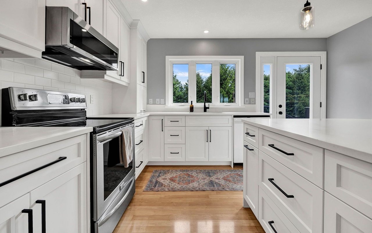 Bright white kitchen with stainless steel appliances, a large island, and windows overlooking trees.
