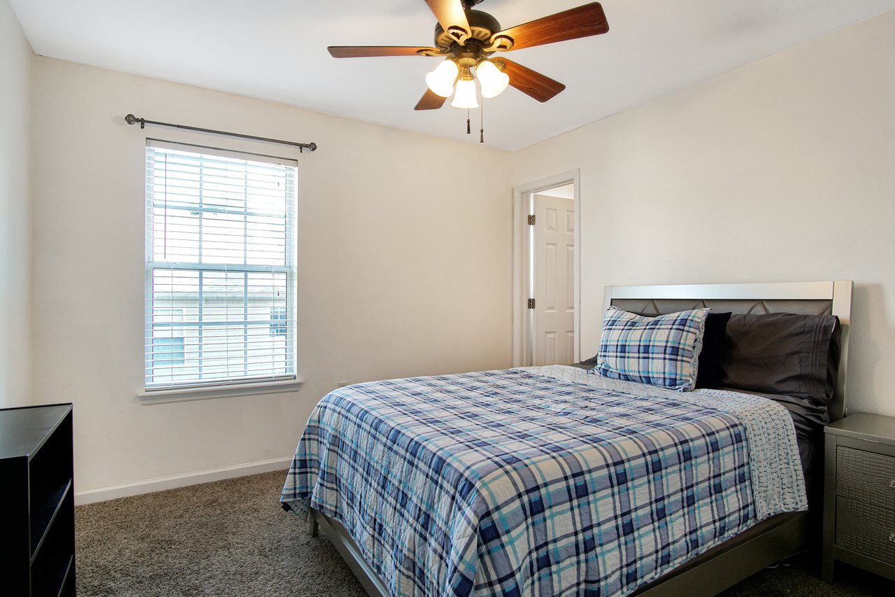 A cozy bedroom features a plaid bedspread on a modern bed, a window with blinds on the left, light beige walls, and a ceiling fan, creating a relaxed vibe.