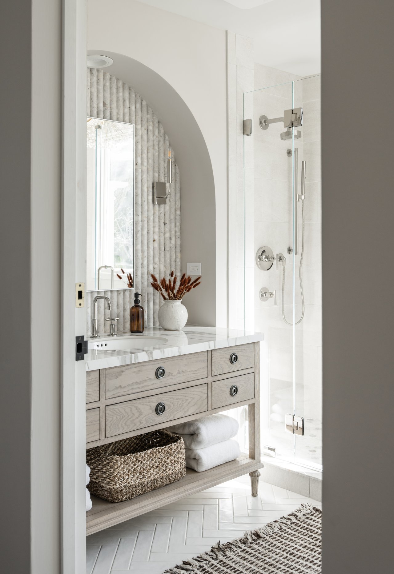Bathroom with a pale wood vanity, arched mirror, and glass-enclosed shower.