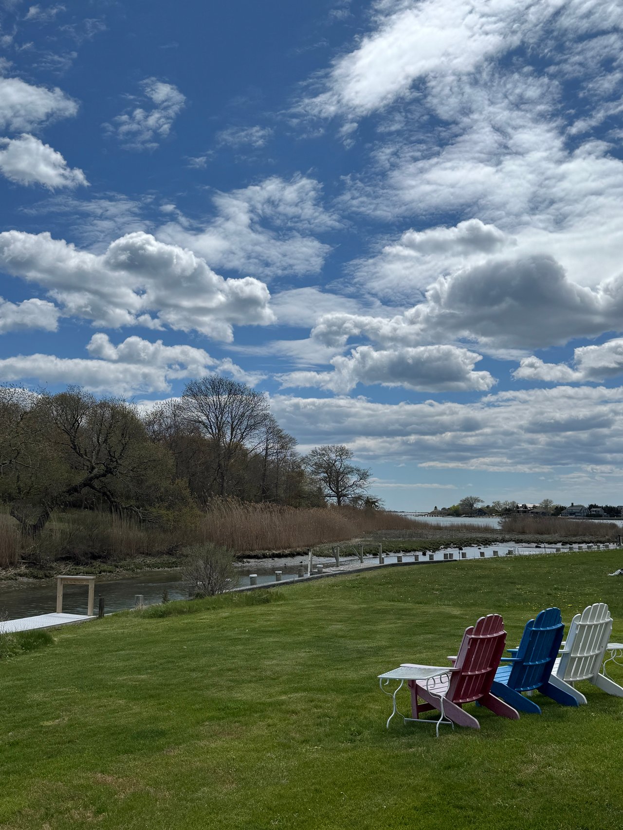 Waterfront lawn with a small dock, creek, and colorful Adirondack chairs beneath a cloud-filled sky.