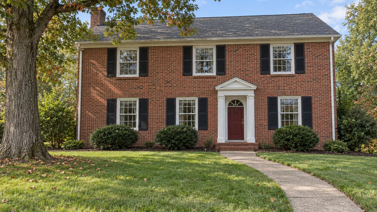 Traditional two-story red brick home with black shutters, a burgundy front door, a deep lawn, and a mature oak.