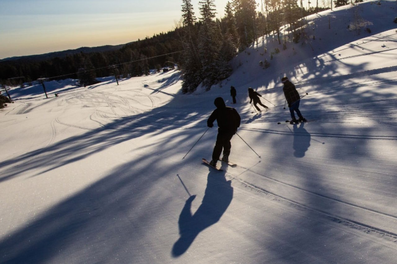Silhouette view of downhill skiers carving down a wide, snow-covered ski trail during a sunny winter morning.