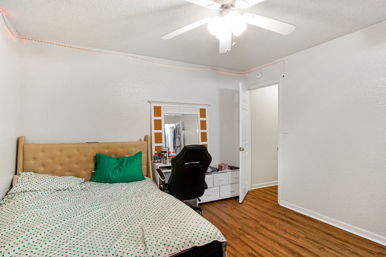Bedroom at 2506 Nugget Lane in Tallahassee, Florida, with wood-look flooring, white walls, and a ceiling fan with light. The room includes a bed with upholstered headboard, a desk with mirror and drawers, and an open doorway leading to the hallway. 