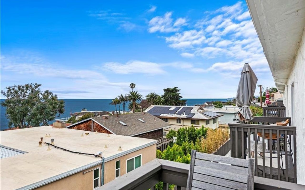 Balcony overlooking the ocean, with palm trees, neighboring rooftops, and outdoor seating.