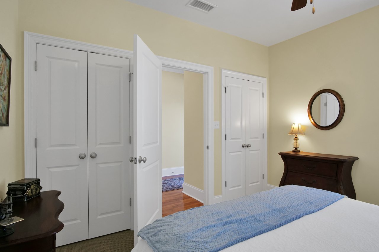 Cozy bedroom with cream walls, a bed with a light blue throw, wooden dresser, round mirror, lamp, closed closets, and an open door to a hallway.