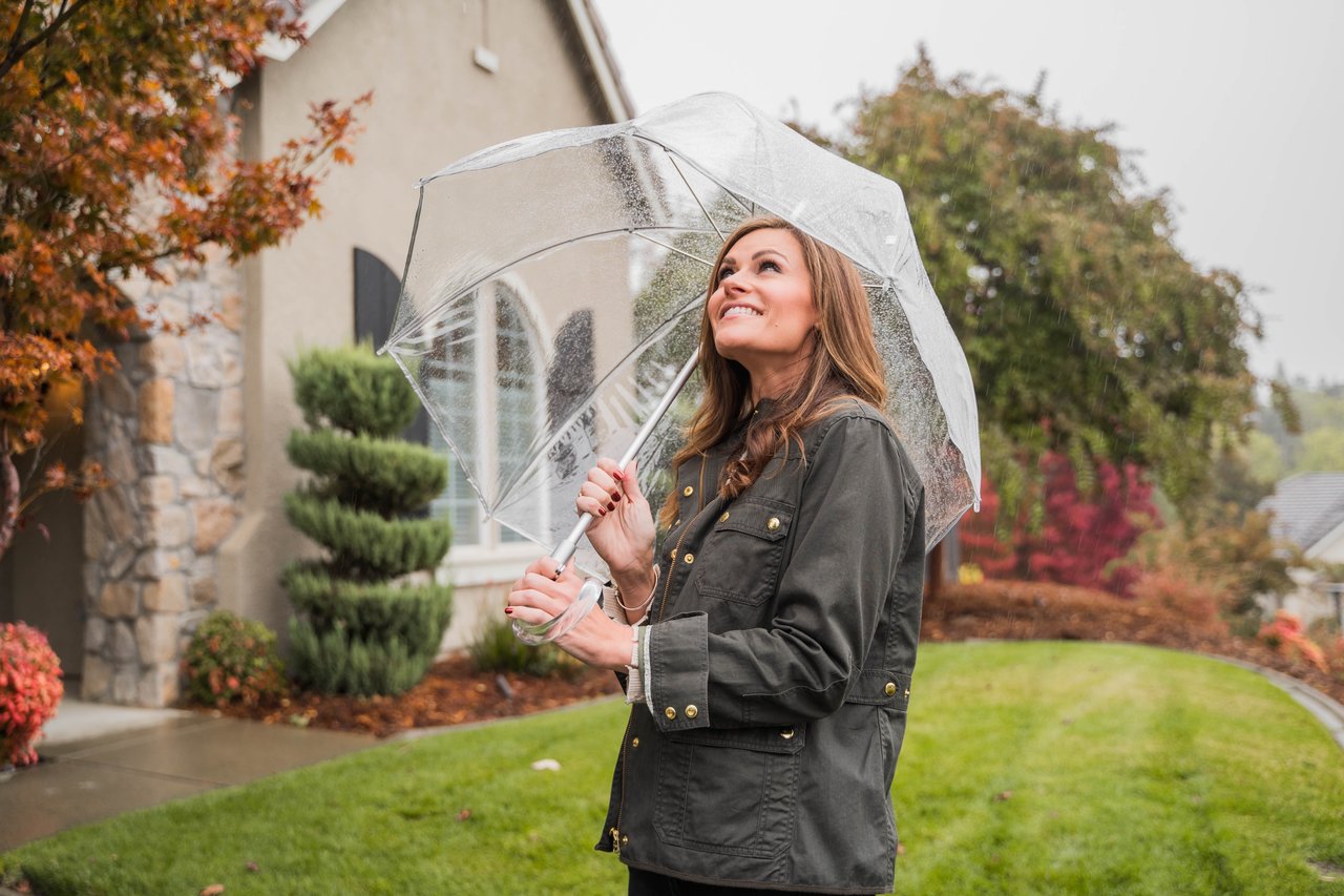 Woman smiling beneath a clear umbrella outside a home on a rainy autumn day.
