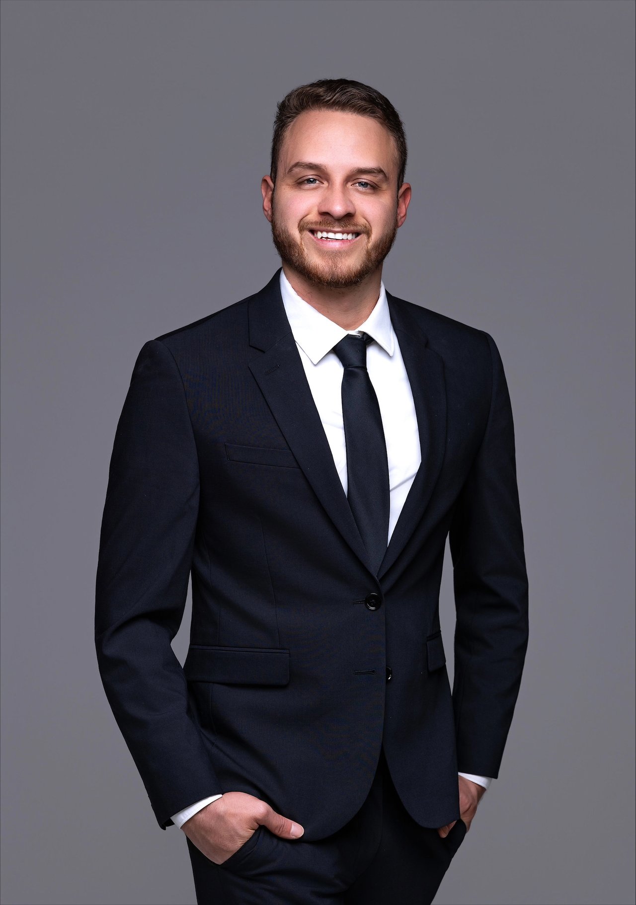 Maykel Orozco-Cruz smiling in a dark suit and tie against a gray background.