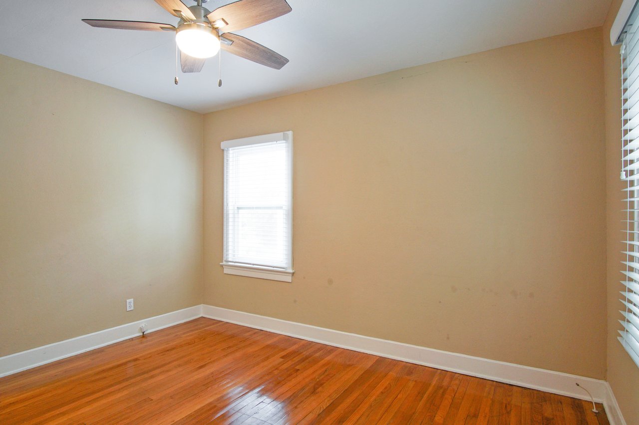 Empty beige room with wooden floor, ceiling fan, and two windows with blinds. Soft natural light creates a warm, welcoming atmosphere.