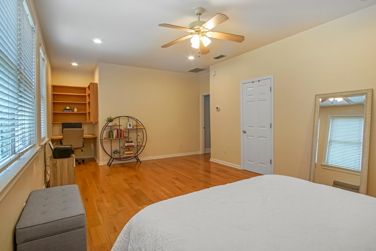 A spacious bedroom with hardwood floors, a ceiling fan, and beige walls, featuring a bed, a large mirror, and a chair. Soft lighting conveys a cozy ambiance.