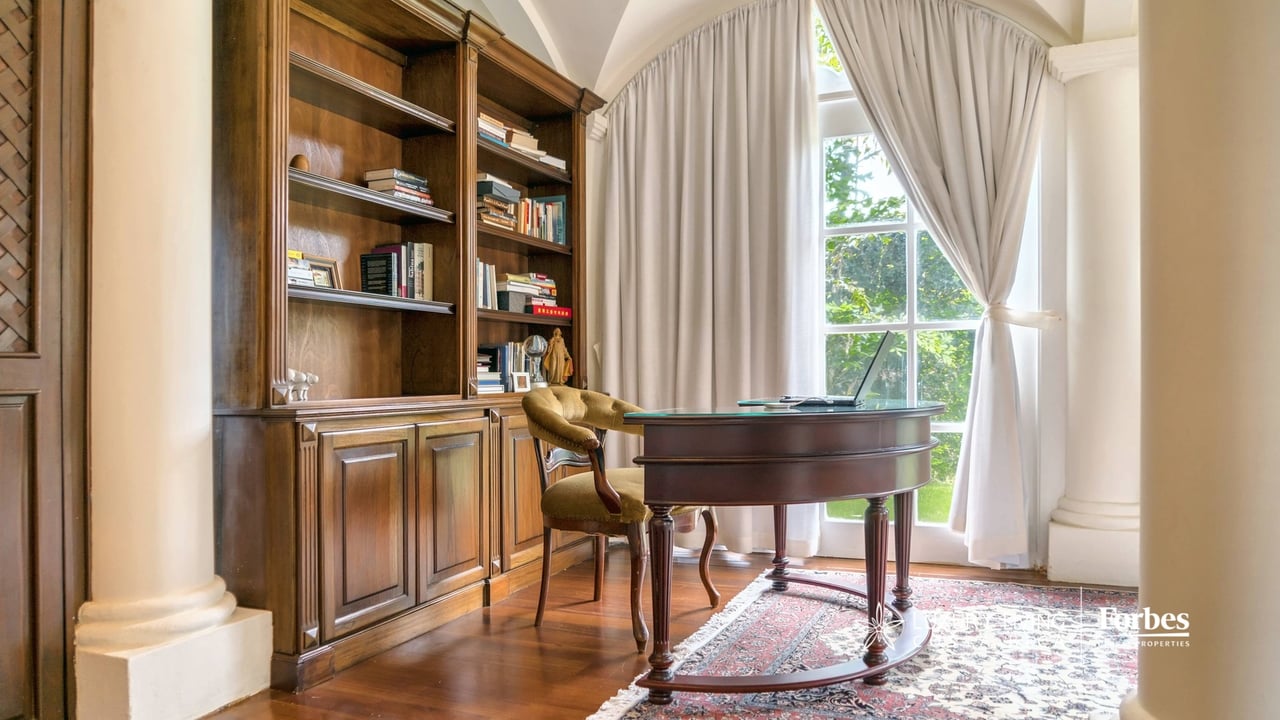 Home study with a curved wooden desk, built-in bookcase, and tall curtained window.