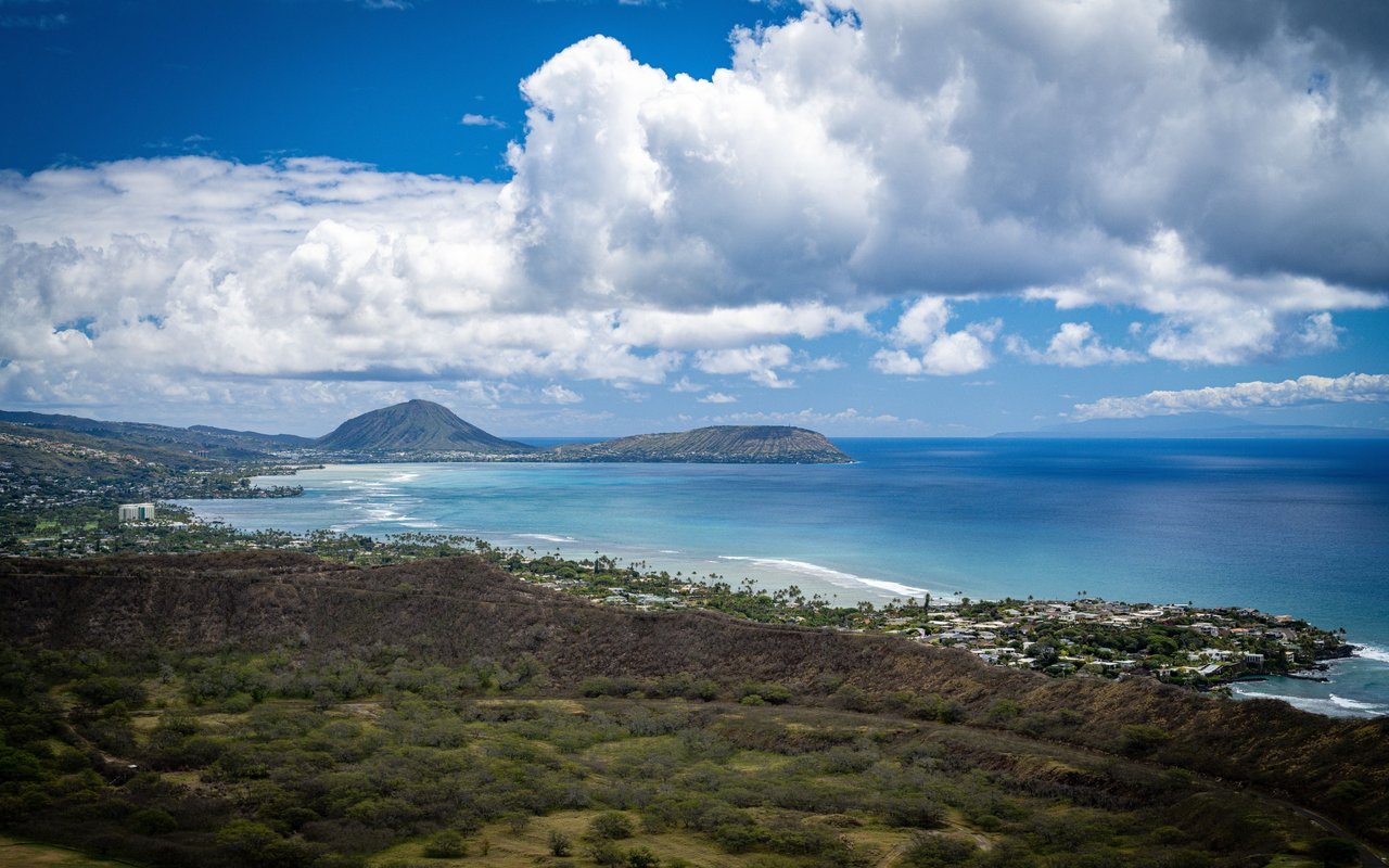 Coastal Oahu and blue ocean viewed from a ridge beneath towering clouds.