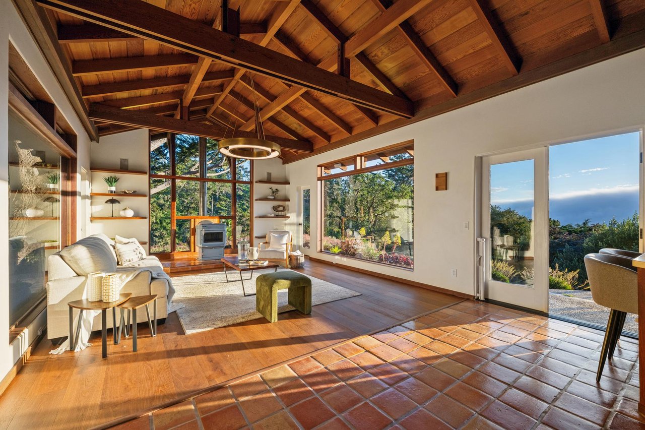 Bright living room with vaulted wood-beamed ceiling, terracotta tile and hardwood floors, large picture windows overlooking forest, and a sliding glass door opening to an ocean view