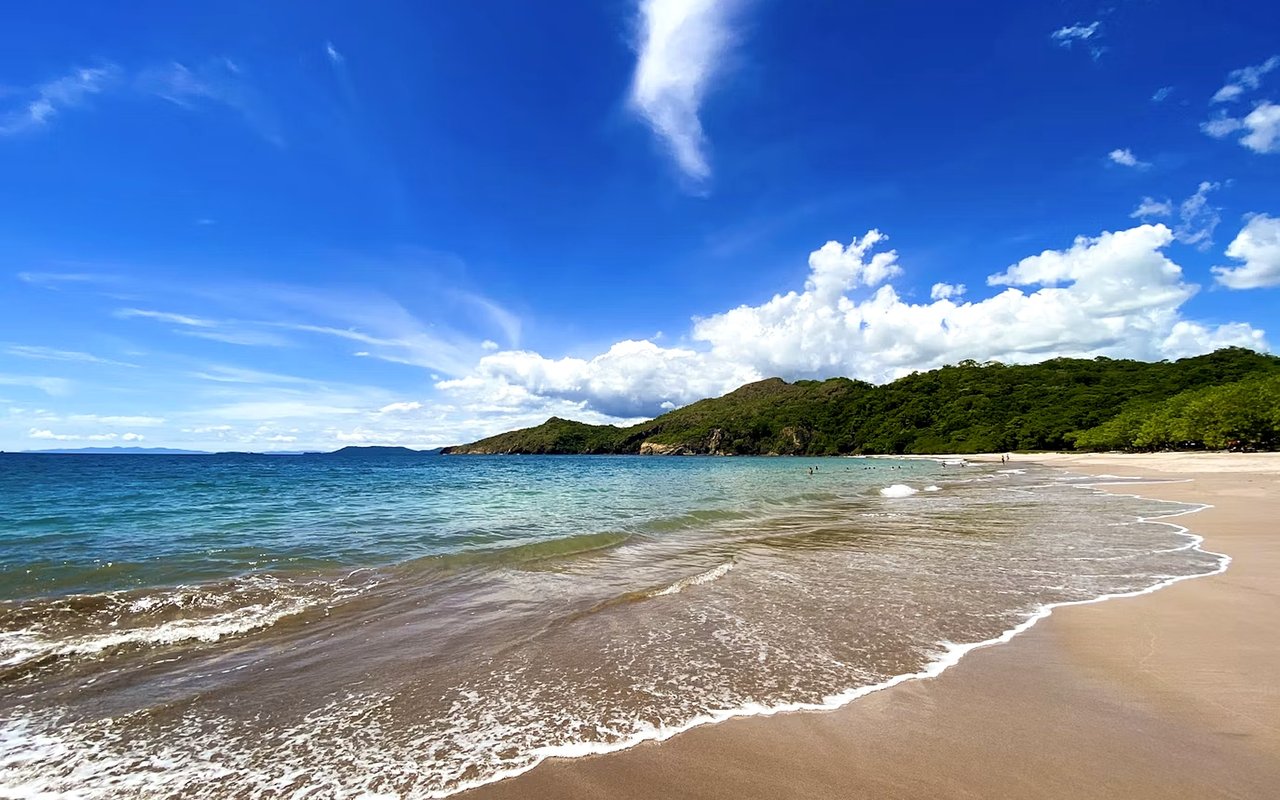 Sandy beach curving beside turquoise water and forested hills under a bright, cloud-filled sky.