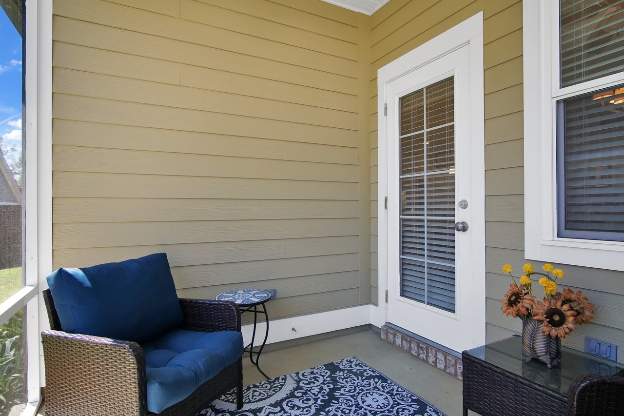 Cozy patio with wicker chair and blue cushion, next to a small table. White French door, floral vase, and decorative rug add a warm, inviting feel.