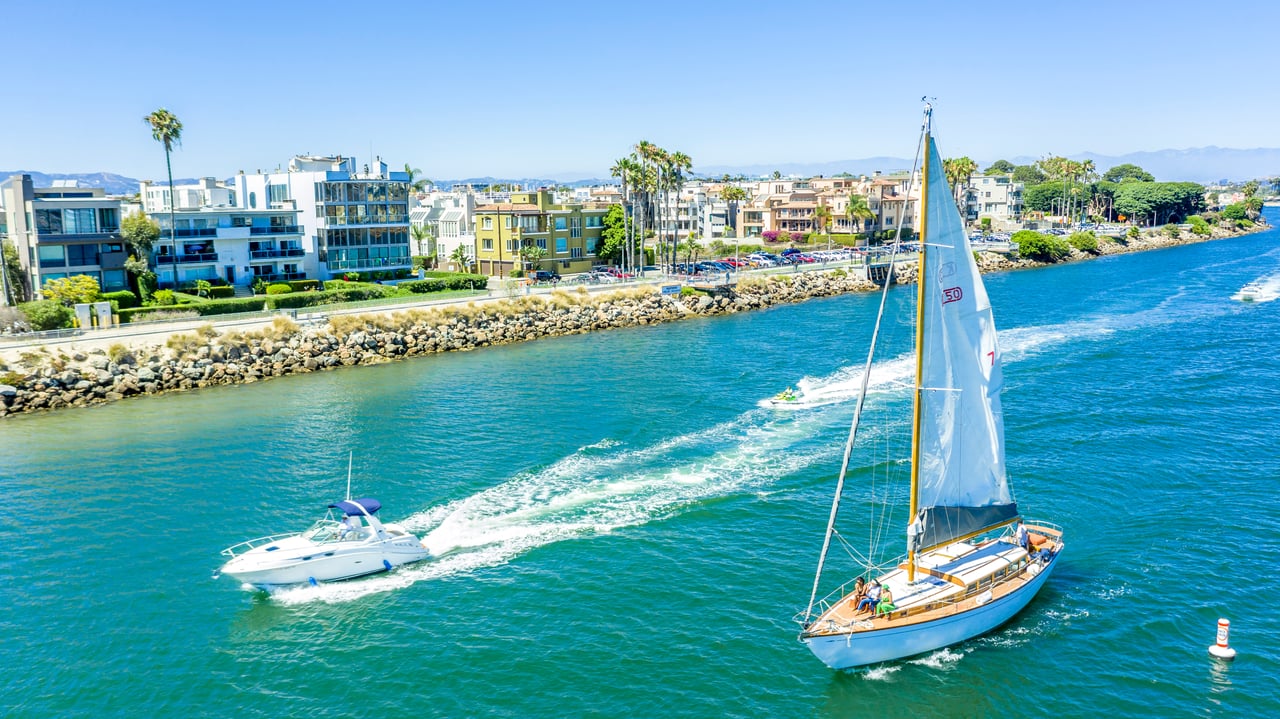 Sailboat and motorboat cruise Marina del Rey’s blue water beside a shoreline of homes and apartments.