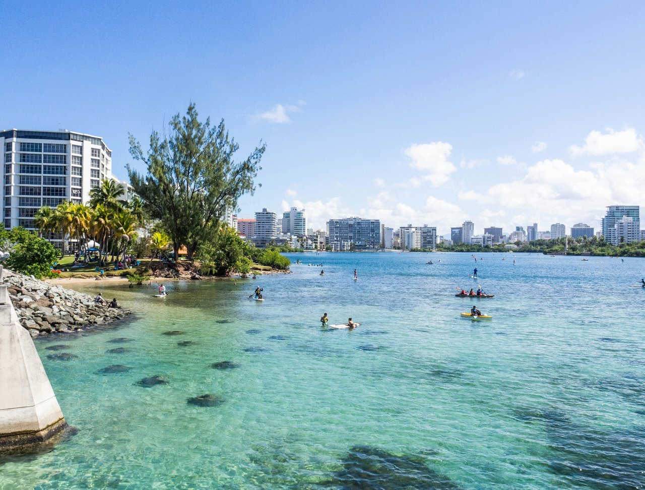 Clear waters of Condado Lagoon with people kayaking and paddleboarding, surrounded by lush greenery and the Miramar skyline in the background.