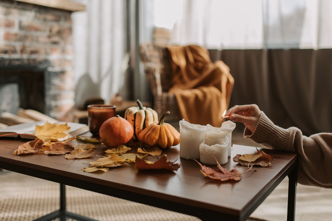 Fall-decorated table with pumpkins and leaves as a person lights candles beside a brick fireplace.