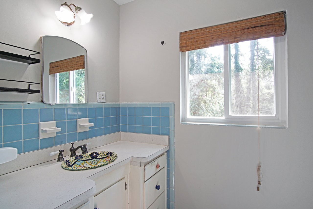 Small bathroom with light blue tiled backsplash, white countertop, and a colorful decorative sink. Natural light filters through a window with a bamboo blind.