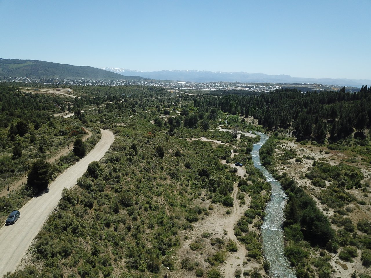 Aerial view of scrubland with a winding dirt road, a tree-lined stream, and distant mountains.