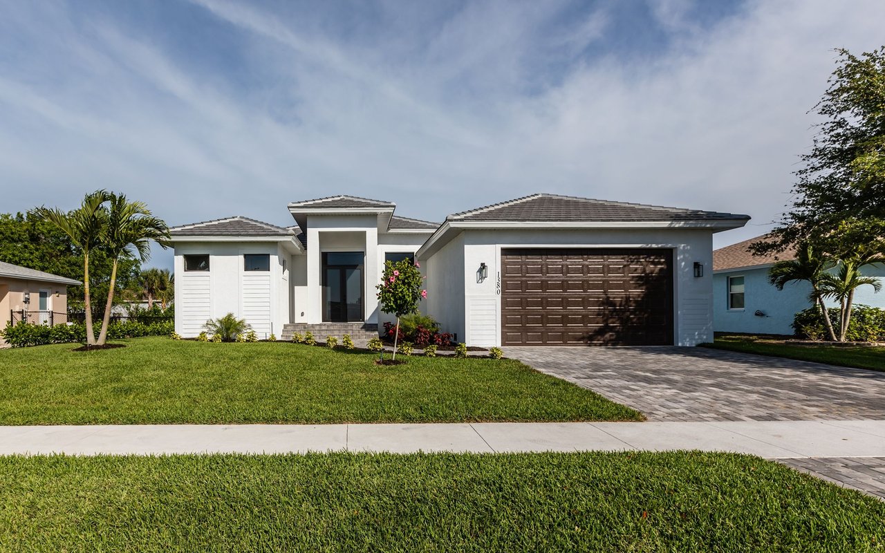 Modern white house with a dark double garage, paver driveway, and palm trees along the front lawn.