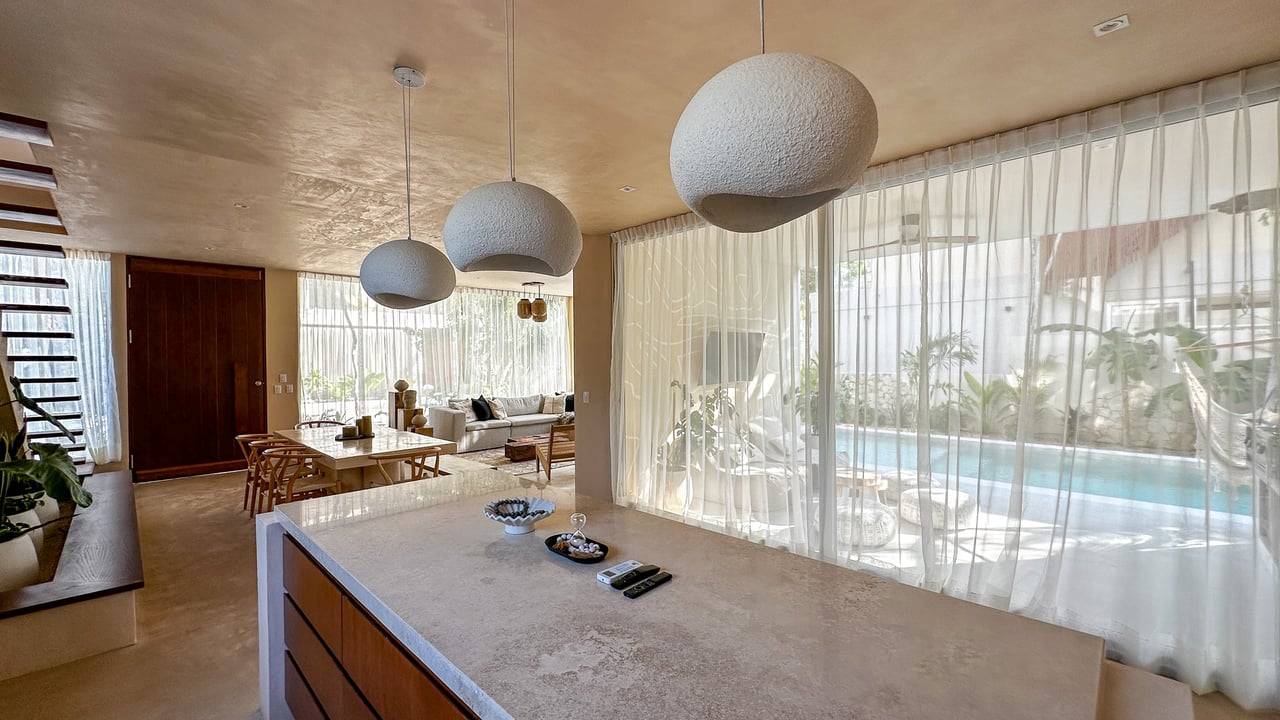 Open-plan kitchen and dining area with sculptural pendant lights and a pool visible through sheer curtains.