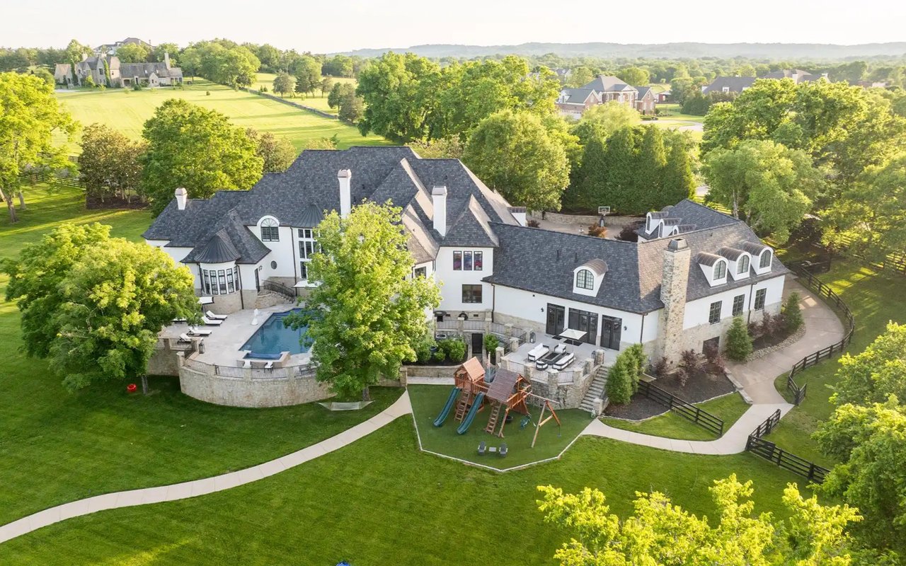 Aerial view of a large white home with a pool, stone terraces, and a children’s playset.