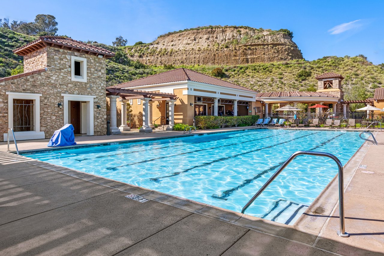 Community swimming pool with lane markings, shaded seating, and hillside buildings.