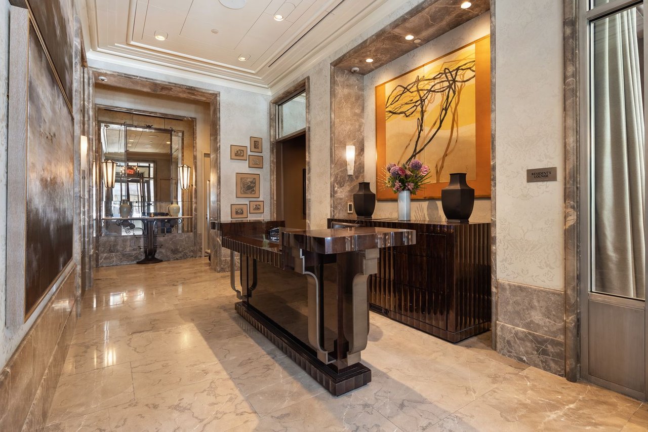 Building lobby with marble floors, a dark wood console, and abstract artwork beside the residents’ lounge.