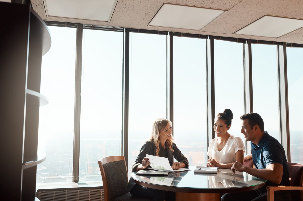 Three real estate agents sit around a table in a high-rise office discussing documents, with large windows showcasing a vibrant cityscape in the background.