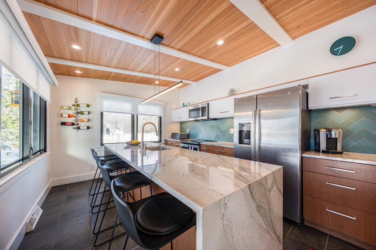 Renovated kitchen with a marble waterfall island, teal tile backsplash, and wood-paneled ceiling.