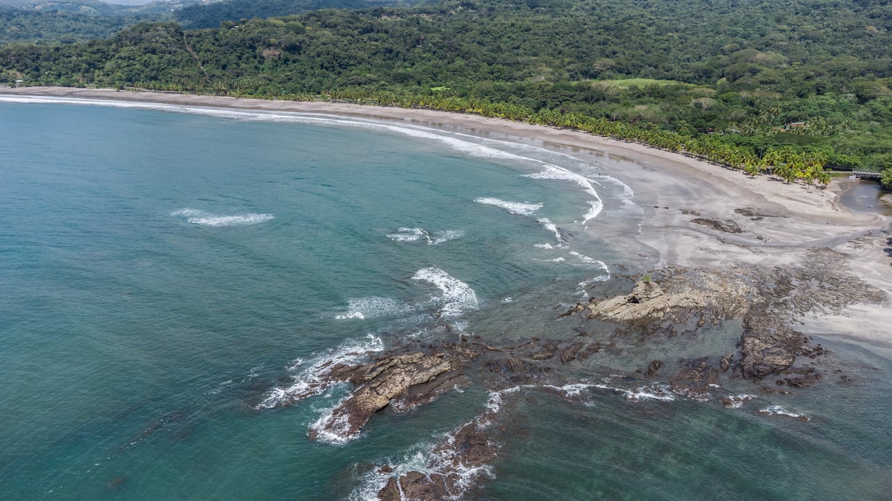 Aerial coastline with a sweeping sandy beach, rocky shoreline, and forested green hills.