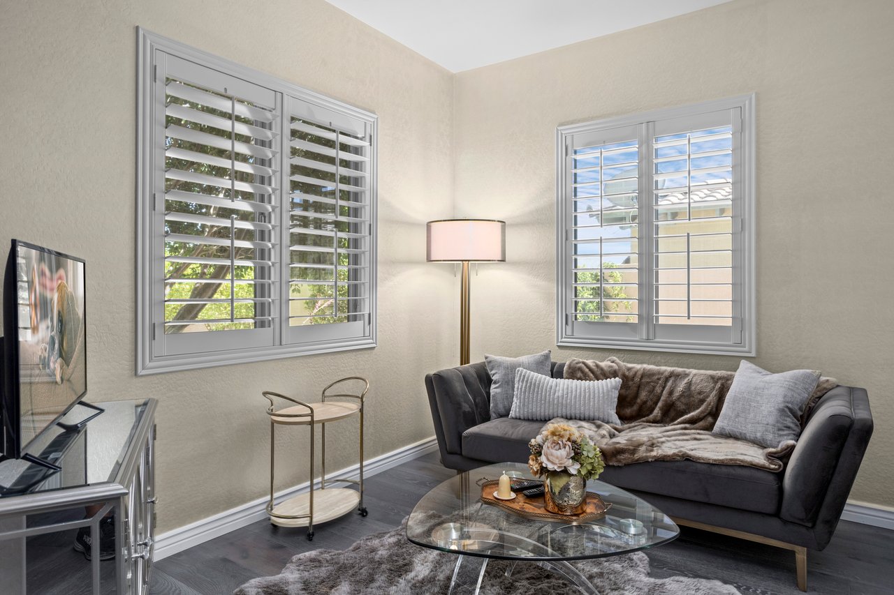 Staged living room corner with a dark sofa, glass coffee table, floor lamp, TV stand, and two windows with white shutters.