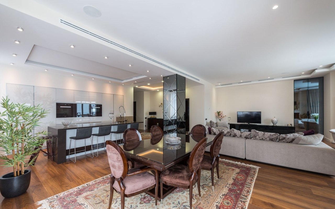 Open-plan living and dining area with a glass table, pale sectional sofa, and modern kitchen.