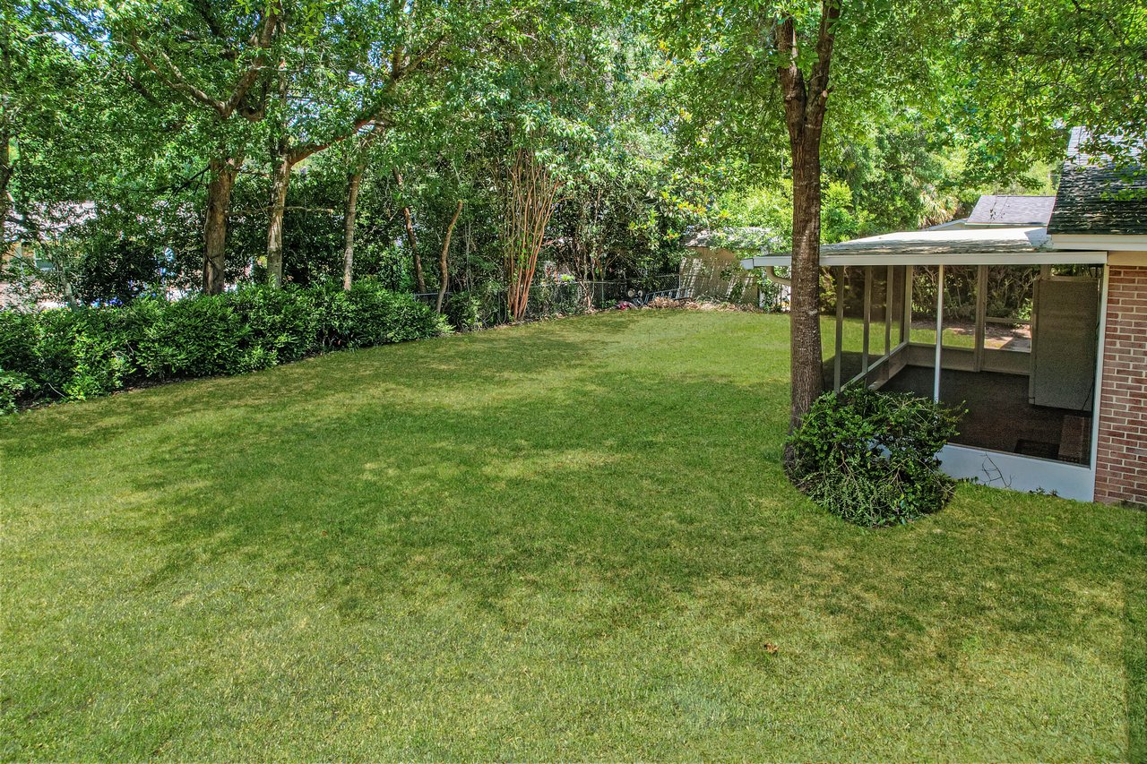 Sunny backyard with lush green grass and tall trees casting shadows. A brick house with a screened porch is on the right, creating a tranquil setting.